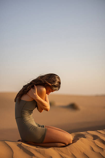 A contemplative woman sits on a sand dune, surrounded by an expansive desert. Her posture conveys solitude and reflection as she embraces herself, with the soft morning sunlight adding warmth to the tranquil atmosphere.