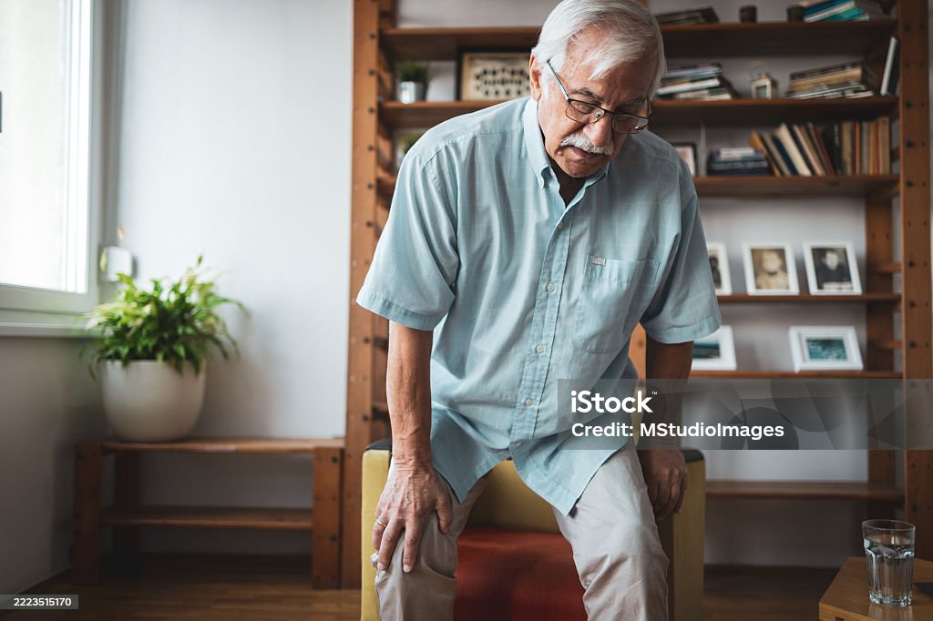 Senior man experiencing joint pain at home Senior adult man holding his knee, experiencing joint pain while standing up in a cozy living room with bookshelves and plants, reflecting on aging and health Chair Stock Photo Senior man experiencing joint pain at home Senior adult man holding his knee, experiencing joint pain while standing up in a cozy living room with bookshelves and plants, reflecting on aging and health Chair Stock Photo