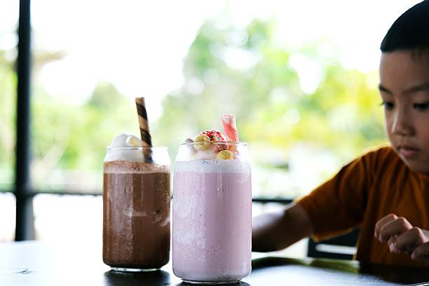 A child excitedly leaning toward a table with chocolate and strawberry milkshakes on a bright day.