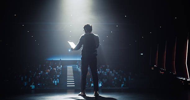 istock Young Empowering Thought Leader Standing Under a Spotlight, Making an Inspiring Presentation About Sustainable Innovations at Technology Conference. Indian Man Talking to an Audience in a Dark Venue 2223521736