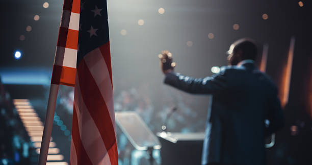 istock Presidential Candidate Meeting with Voters Before Elections and Making a Heartwarming Speech. Stage with an American Flag and Dramatic Lighting, Diverse Blurred Audience Sitting in the Background 2223522856