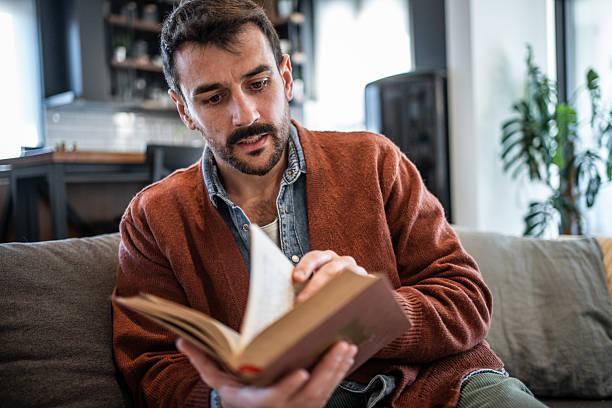 istock Young man reading a book on the sofa at home 2223527232
