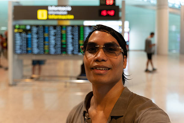 Portrait of a happy young adult man taking a selfie in front of the flight information display in an airport terminal