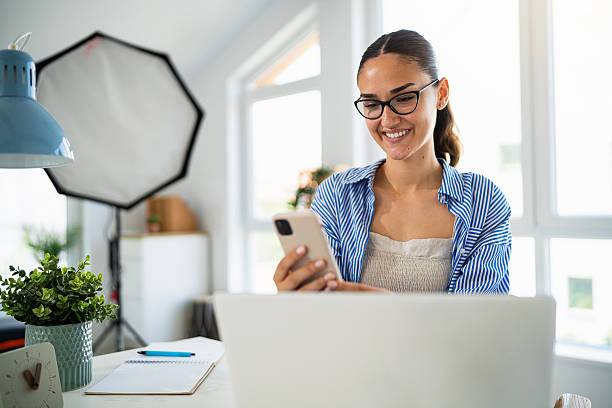 istock Smiling young female photographer using smartphone while working with laptop in bright modern office 2223531498