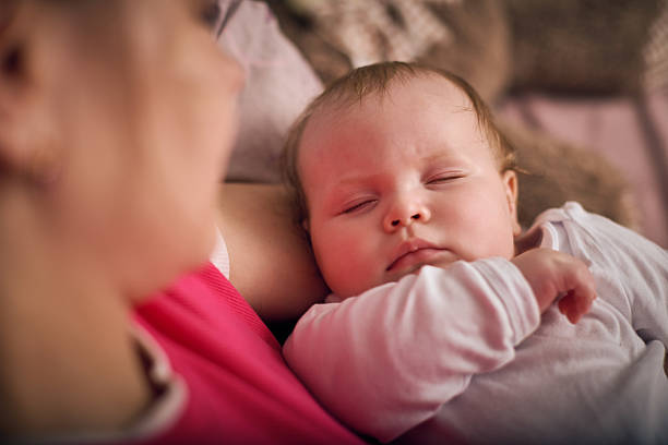 Young blonde woman cradles sleeping baby girl with light hair. Warm lighting, soft background create comforting atmosphere. Pink, beige hues dominate scene