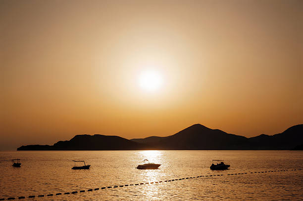 Silhouetted boats on a calm sea at sunset with mountains in the background.