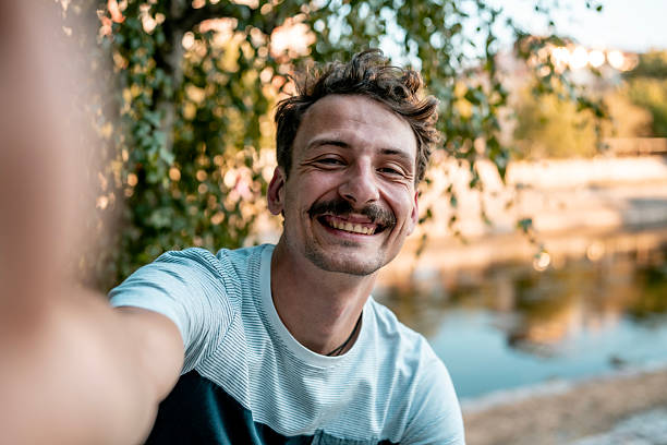 Happy man smiling brightly while taking a selfie in an outdoor setting with lush greenery and a scenic lake in the background, conveying joy and relaxation in nature.