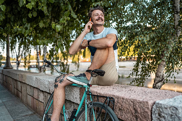 istock Smiling Young Man Talking on Phone While Relaxing with Bicycle Outdoors 2223551129
