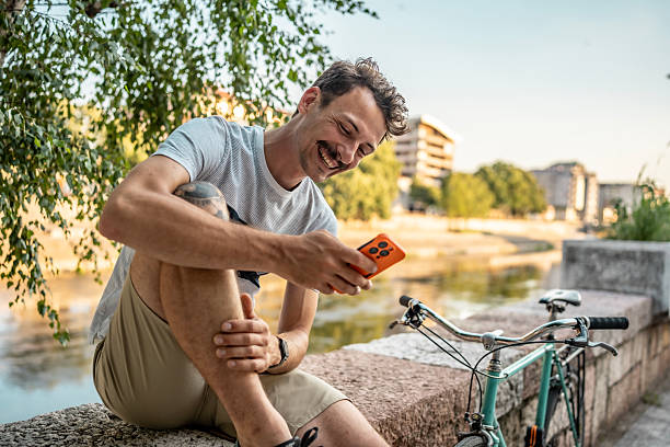 istock Man Smiling by a Bicycle Using Phone Seated Near a River 2223551176