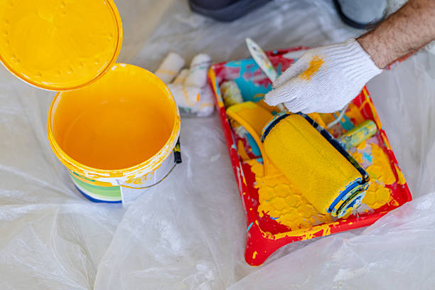 A house painter applies yellow paint to a wall using a paint roller. The painter is focused and dressed in professional attire while surrounded by paint supplies in a spacious indoor area.