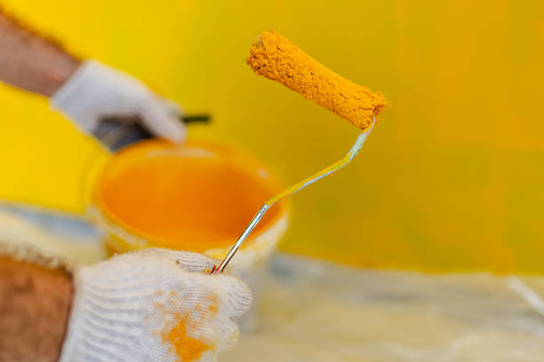 A house painter focuses on applying vibrant yellow paint to a wall with a roller. The painter wears protective gloves and works indoors, improving the space's appearance.