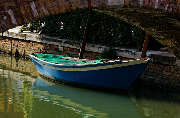 Burano, Italy - June 25, 2025: Blue boat is anchored under a brick bridge, with tranquil water and lush greenery creating a peaceful scene in the canal