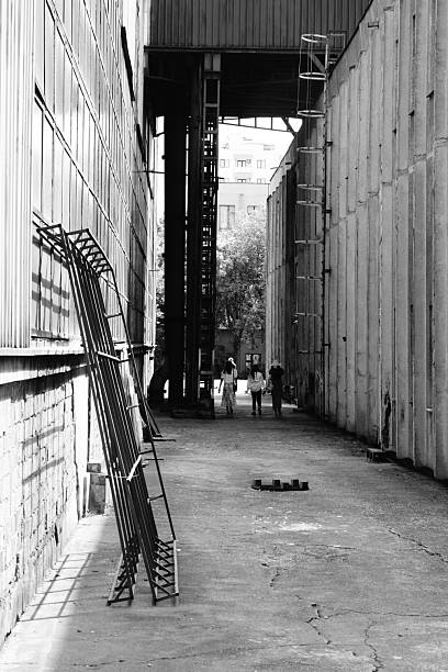 A stack of metal ladders leans against a wall in a long, narrow alleyway framed by high, textured walls and figures walking away in the distance.