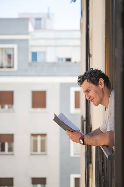 Mature man enjoying a book on his balcony, soaking in the city view and fresh air while embracing a moment of relaxation and peace