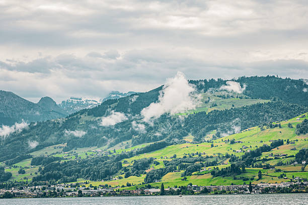 Panoramic view of Swiss Alpine foothills with vibrant green pastures, small town on the lakeshore, and misty clouds drifting across the forested mountains. Captured in Switzerland.