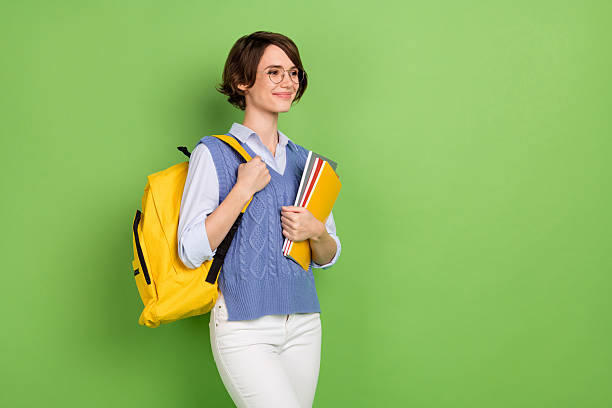 istock Photo portrait of female student with yellow rucksack keeping book stack looking copyspace isolated bright green color background 2223578152