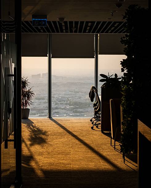 Riyadh, Saudi Arabia – August 11, 2023: Office interior with floor-to-ceiling windows, plants, and city view at sunset, casting long shadows on the carpeted floor.