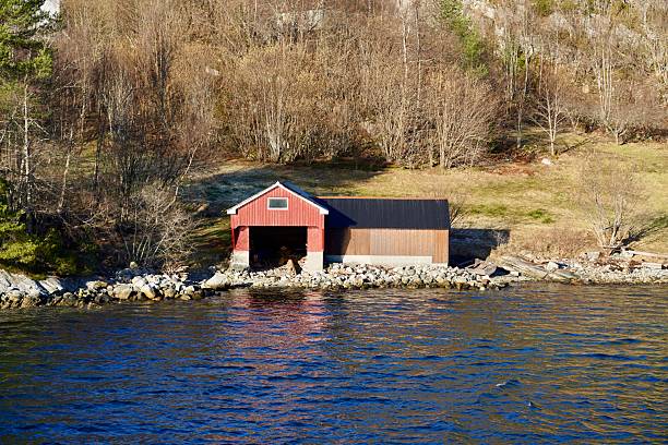 A red painted boathouse stand on the shore of a Norwegian fjord.