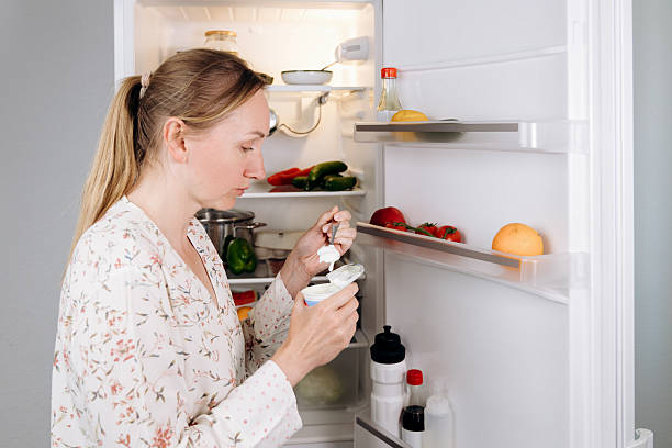 istock Woman enjoying yogurt from a container while standing in front of an open refrigerator filled with fresh fruits and vegetables, emphasizing healthy dairy alternatives in a modern kitchen setting 2223599738