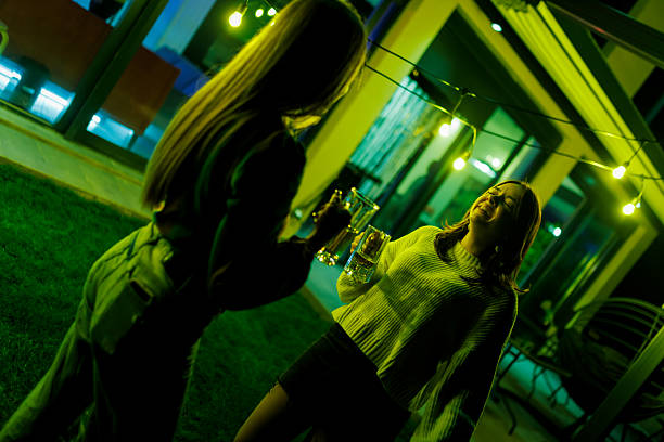 Two women share drinks during a vibrant evening gathering, illuminated by warm string lights. The celebration takes place outdoors, reflecting friendship, joy, and leisure time spent together.