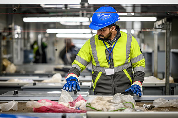 A diligent worker in a high-visibility jacket and blue helmet meticulously inspects materials on a conveyor belt, embodying the essence of, captured with a three-quarter length portrait, quality control in a recycling center. His focused demeanor underscores the critical role of responsible waste management.