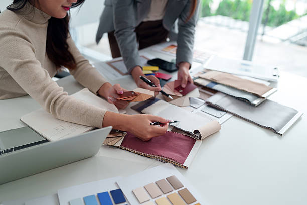 Two adult Asian women interior designers are collaborating on a design project selecting fabrics and colors for a home interior, working with swatches in design studio.