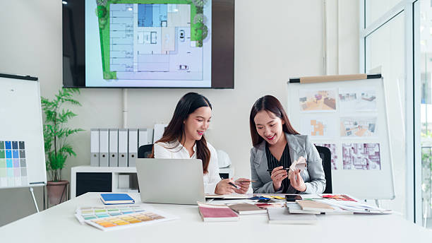 istock Asian Women Interior Designers Collaborate on Home Renovation Project Using Color Palettes and Laptop for Design Planning in Bright Modern Office 2223614359