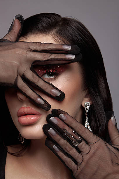woman poses elegantly with her hands partially covering her face. Her striking makeup features bold colors. She wears stylish gloves and accessories, showcasing a modern fashion statement