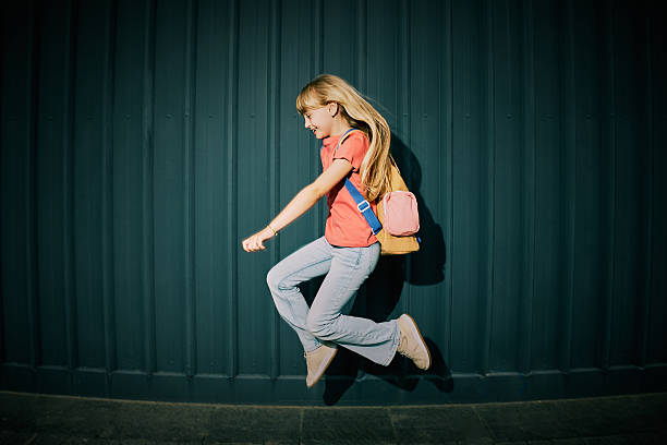 A cheerful blonde schoolgirl with long hair jumps energetically outdoors. She wears casual attire and a pink backpack, set against a modern, dark metal wall under bright sunlight.