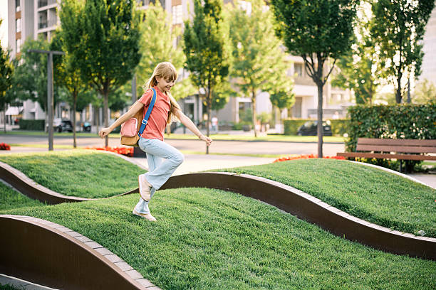 A cheerful young blonde girl wearing backpack enjoys playing on a wavy grass lawn in a sunny urban park. The background features trees, benches, and modern buildings.