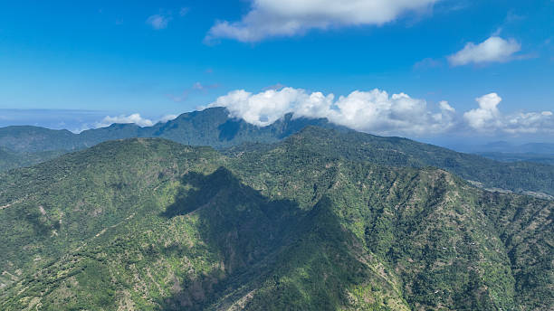 Rolling green mountains stretch into the distance as fluffy clouds drift across the summits. The interplay of sunlight and shadow reveals the wild beauty of this untouched tropical paradise