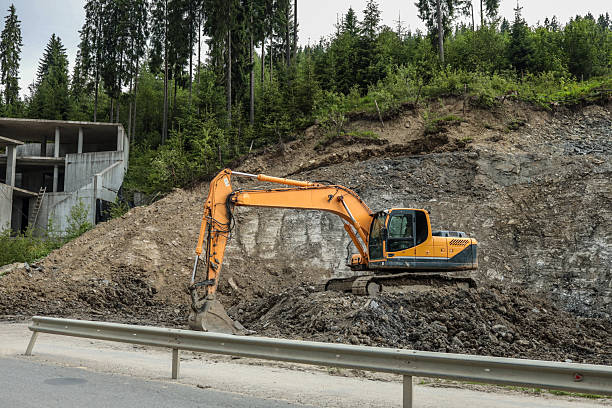 istock Excavator working at construction site near concrete building structure. Heavy machinery digging on rocky slope by the road with forest trees in background 2223630974