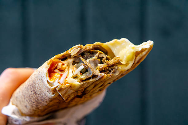 istock hand holding a wrap kebab with visible meat, vegetables, and melted cheese. Dark blurred background contrasts with the appetizing food. 2223632795