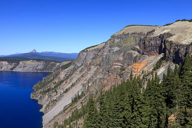 A view of Crater Lake in southern Oregon, with the colorful cliffs of Pumice Castle in the foreground and the sharp peak of Mount Thielsen rising in the distance beyond the caldera rim.