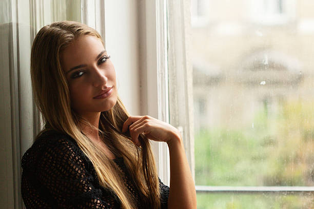 A young woman with long blonde hair sits gracefully by a bright window, wearing a black lace top while gazing thoughtfully with her hand gently touching her hair. The soft natural light filtering through the window creates an atmosphere of tranquility and introspection, symbolizing the beauty found in quiet moments of self-reflection and inner peace.