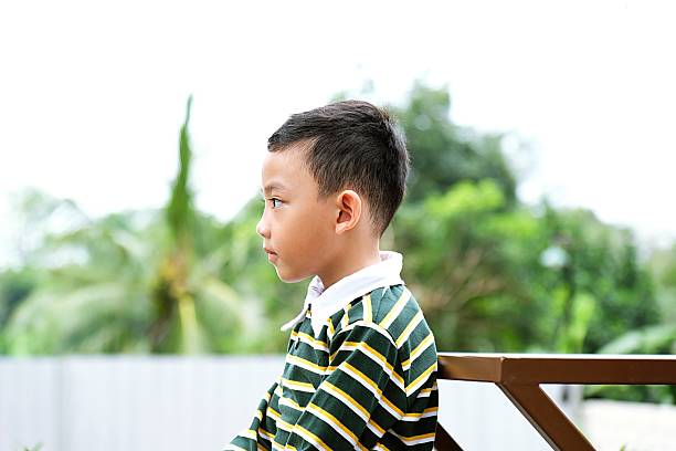 A young boy in a striped shirt sitting sideways outdoors, looking thoughtful.