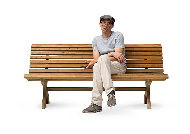 istock Bored mature man in his 50s sitting on a wooden bench and using a smartphone while looking at the camera. Studio shot, isolated on white background. Casual clothing, wearing reading glasses and a hat 2223654080