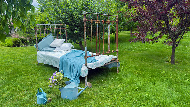 A delightful outdoor snapshot showing a vintage bed adorned with soft pillows and blankets, complemented by a watering can and vibrant flowers, set in a serene garden.