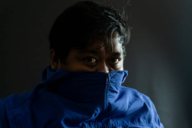 Bearded Indonesian male wearing blue nylon jacket with partially covered face, low-key portrait suggesting secrecy and urban fashion on black backdrop