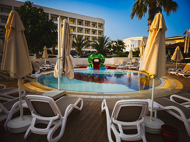 Editorial image showing the children's pool area at Salamis Bay Conti Hotel in Northern Cyprus. The circular pool is surrounded by white loungers and closed umbrellas, with a green dragon-themed slide in the center. Captured during golden hour on June 15, 2025.