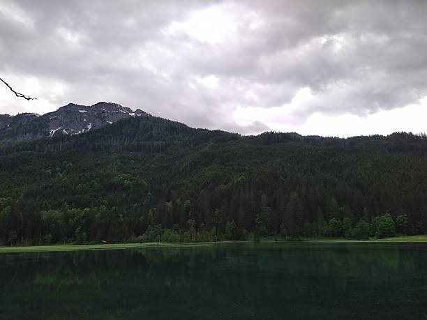 A tranquil mountain lake reflects the rich green hues of a dense evergreen forest and the rugged slopes beyond. Above, thick gray clouds hang low over the landscape, adding a dramatic mood to the serene natural setting. The stillness of the water and the lush surroundings highlight the untouched beauty of this mountainous wilderness, ideal for themes of relaxation, adventure, and nature appreciation.