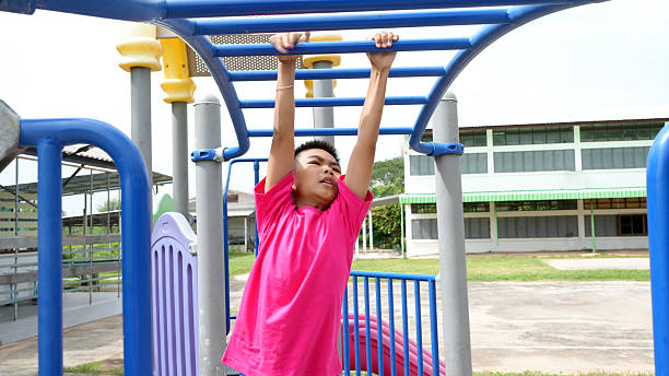 istock Young boy in pink shirt is hanging from playground structure, showcasing his strength and determination. playground is set in school environment, with buildings in background 2223680218