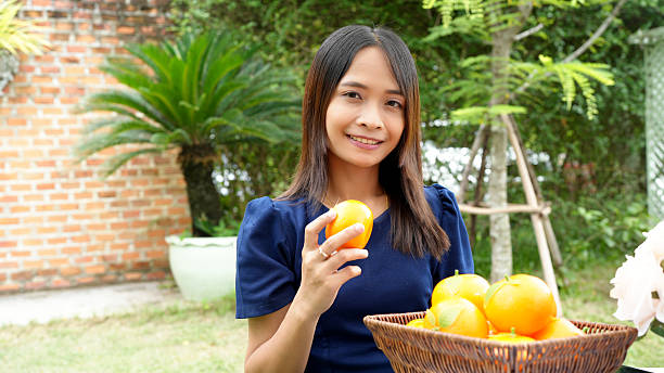 A young woman smiles while holding orange in garden setting, surrounded by greenery and basket of oranges. scene conveys sense of joy and freshness