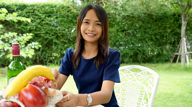 Woman sitting at table outdoors, smiling and holding bowl of fresh fruits. setting is lush and green, creating cheerful atmosphere
