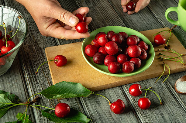 istock Hands carefully sort shiny red cherries on a kitchen table. A green bowl holds fresh cherries while leaves and sugar accompany them. The setting suggests a rustic fruit preparation 2223685249