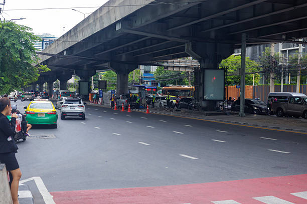 Thai construction workers below overpass road at Rama 9 Road in Bangkok. Workers are unloading equipment from cars. At left side a woman is standing at stoplight waiting for green signal to cross the street. Behind elevated road many cars are queued on opposite lanes in front of stoplight and junction ahead