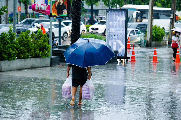 Rear view of barefoot walking thai man in hard rain in Bangkok. Scene is at Ratchadaphisek Road in business area between Street 23 and Esplanade building. man is walking and has a large umbrella. With both hands he is carrying heavy bags. He is passing towards driveway and sidewalk at late afternoon in rainy season.  On street is rush hour traffic