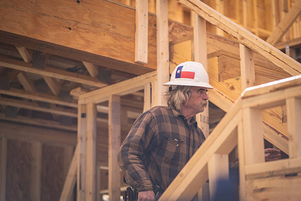 A bearded male construction worker in grey overalls and red plaid shirt holds a red helmet inside a wooden house under construction, surrounded by timber and building tools, representing active building work indoors