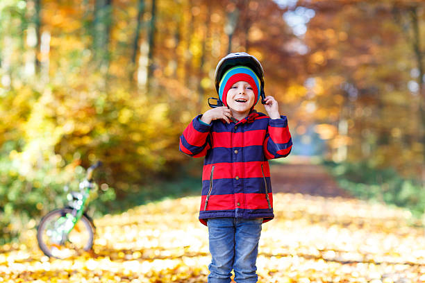 Little kid boy in colorful warm clothes in autumn forest park with a bicycle. Active child putting safe helmet before cycling on sunny fall day in nature. Safety, sports, leisure with kids concept.