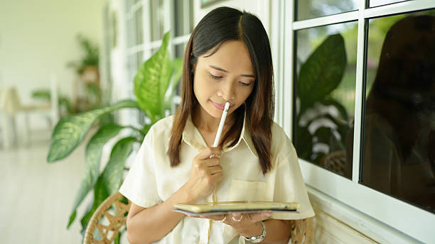 Woman is thoughtfully writing in notebook while sitting by window surrounded by greenery. serene atmosphere enhances her focus and creativity